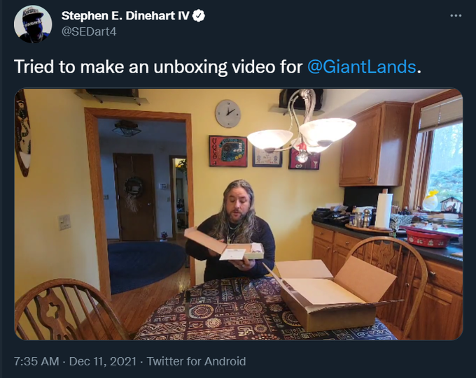 A screencap of a tweet by Stephen E. Dinehart IV (verified) @SEDart4
"Tried to make an unboxing for @GiantLands"
below is a photo of Dinehart in a kitchen with a box, and him looking into the box rather than showing the content - his hair is messily spread across his shoulders.