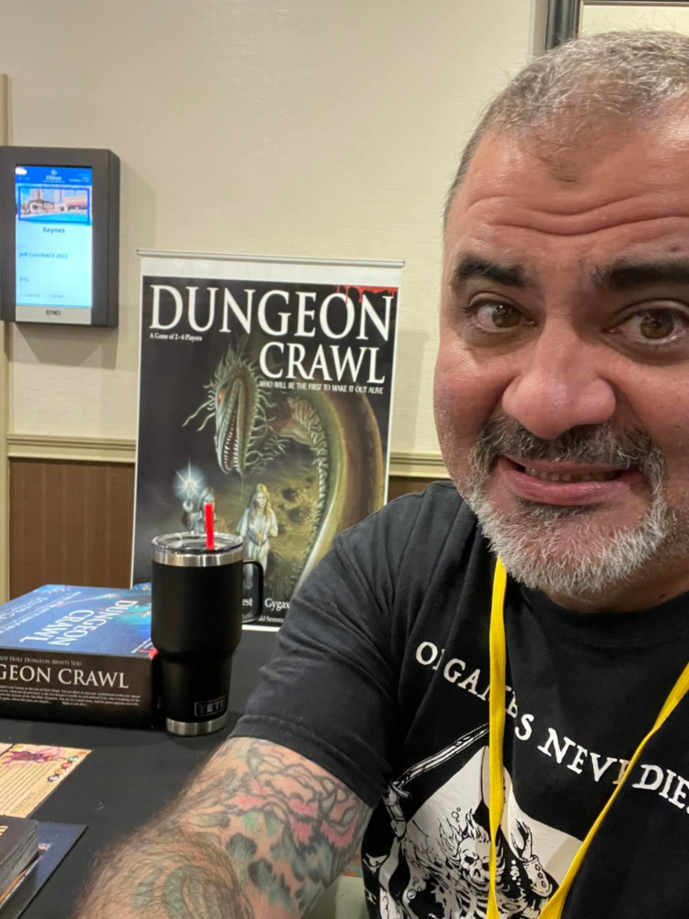 A selfie taken by Justin LaNasa, attempting to showcase his stand at JeffCon, which has very minimal display for Dungeon Crawl and appears to be a very small table.  Justin himself looks uncertain and embarrassed as he takes the the selfie.