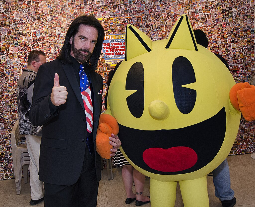 A photo of Billy Mitchell, a middle-aged white man with a beard and shoulder length hair, in a black-on-black suit with an American tie flag, standing and giving the thumbs up, while holding hands with a Pacman mascot.