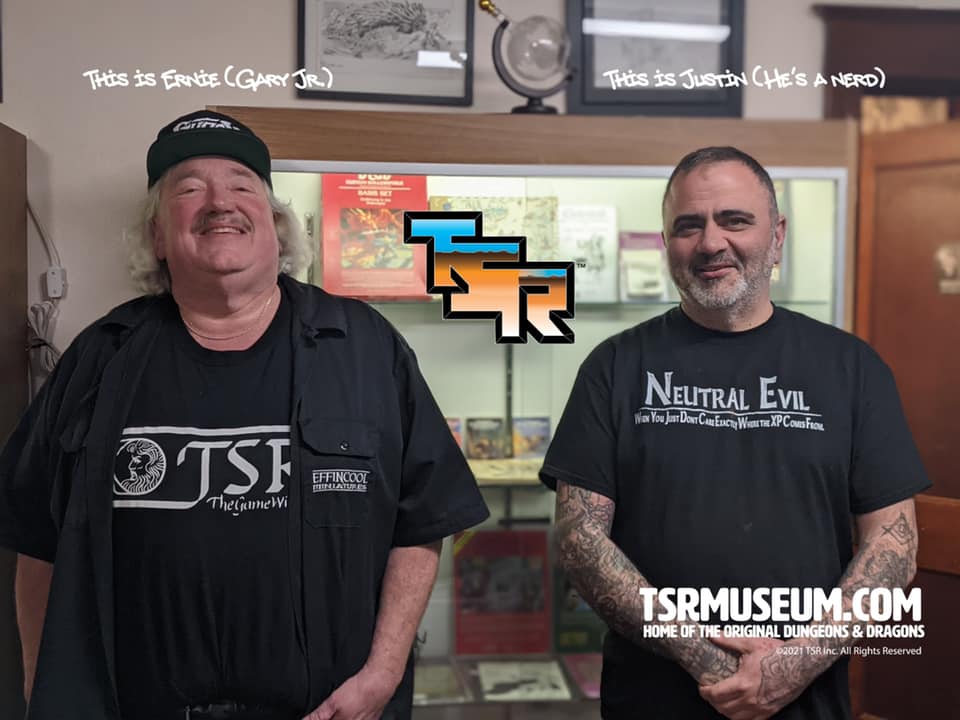 A photo of Ernie Gygax standing next to Justin LaNasa, from the start of the fiasco, with the weird Transformers type hybrid logo. Justin is wearing a "Neutral Evil" "When You Just Don't Care Exactly Where the XP Comes From" t-shirt.